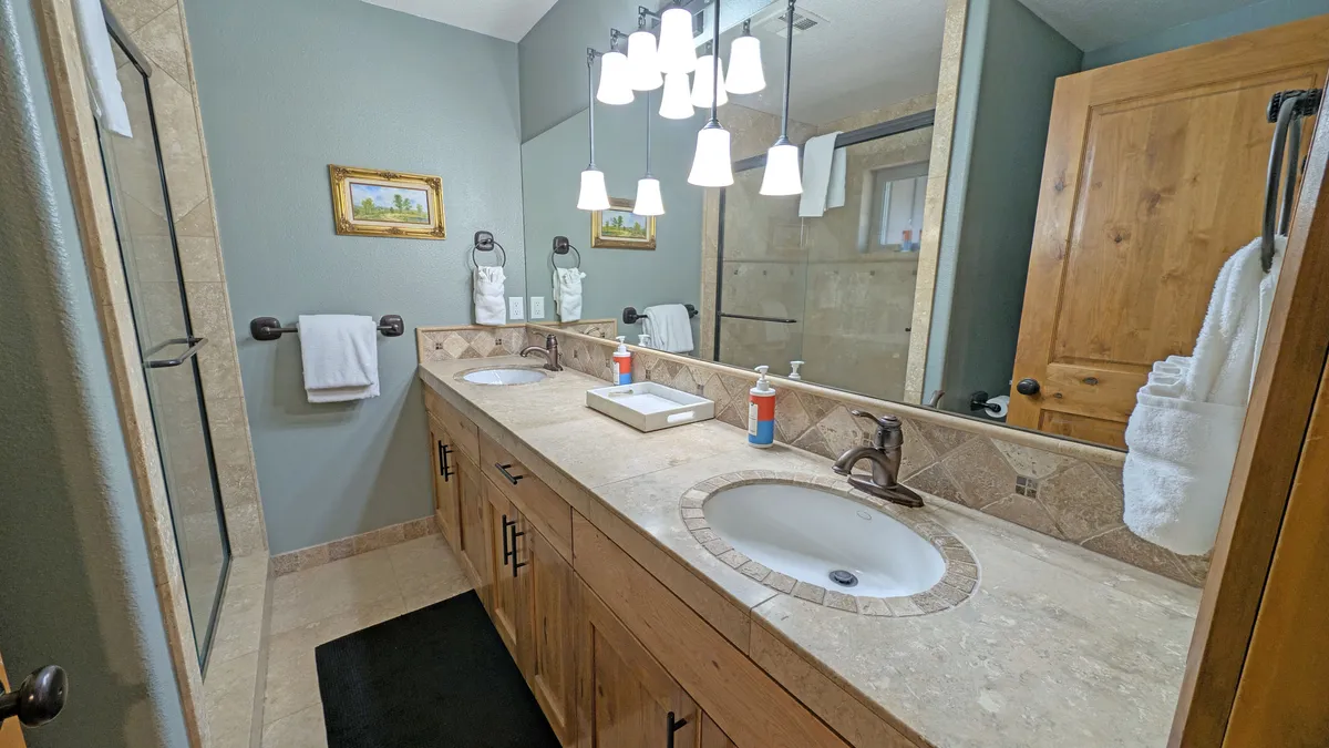 A serene en suite bathroom with a double sink vanity, framed artwork, and elegant chandelier lighting. The room is bright, offering a spa-like atmosphere ideal for relaxation and self-care routines.