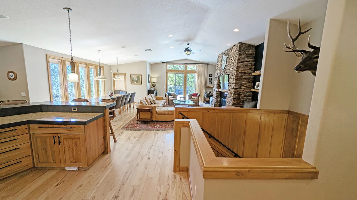 This image showcases a spacious open-concept living area featuring a large kitchen island, adjacent dining space with a sizable wooden table, comfortable living room seating, and a stone fireplace. The area is illuminated with natural light from oversized windows, creating a warm and inviting atmosphere ideal for gatherings.