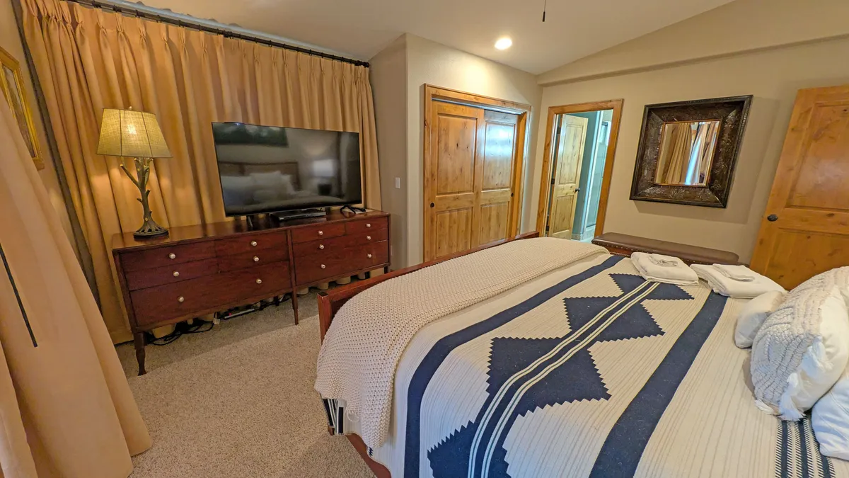 A king bedroom with a patterned blanket on the bed, a wooden dresser, a television, a framed mirror, and curtains. The space feels tranquil and well-appointed, with ample natural light and modern amenities visible.