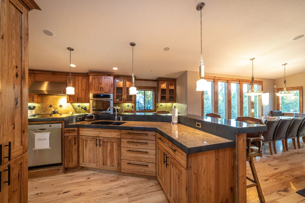 This image showcases a bright, spacious kitchen featuring a large kitchen island with wood cabinetry, modern stainless steel appliances, and an adjacent dining area with ample natural light streaming through large windows. The setup is ideal for meal prep and group gatherings, highlighting generous counter space and seating.