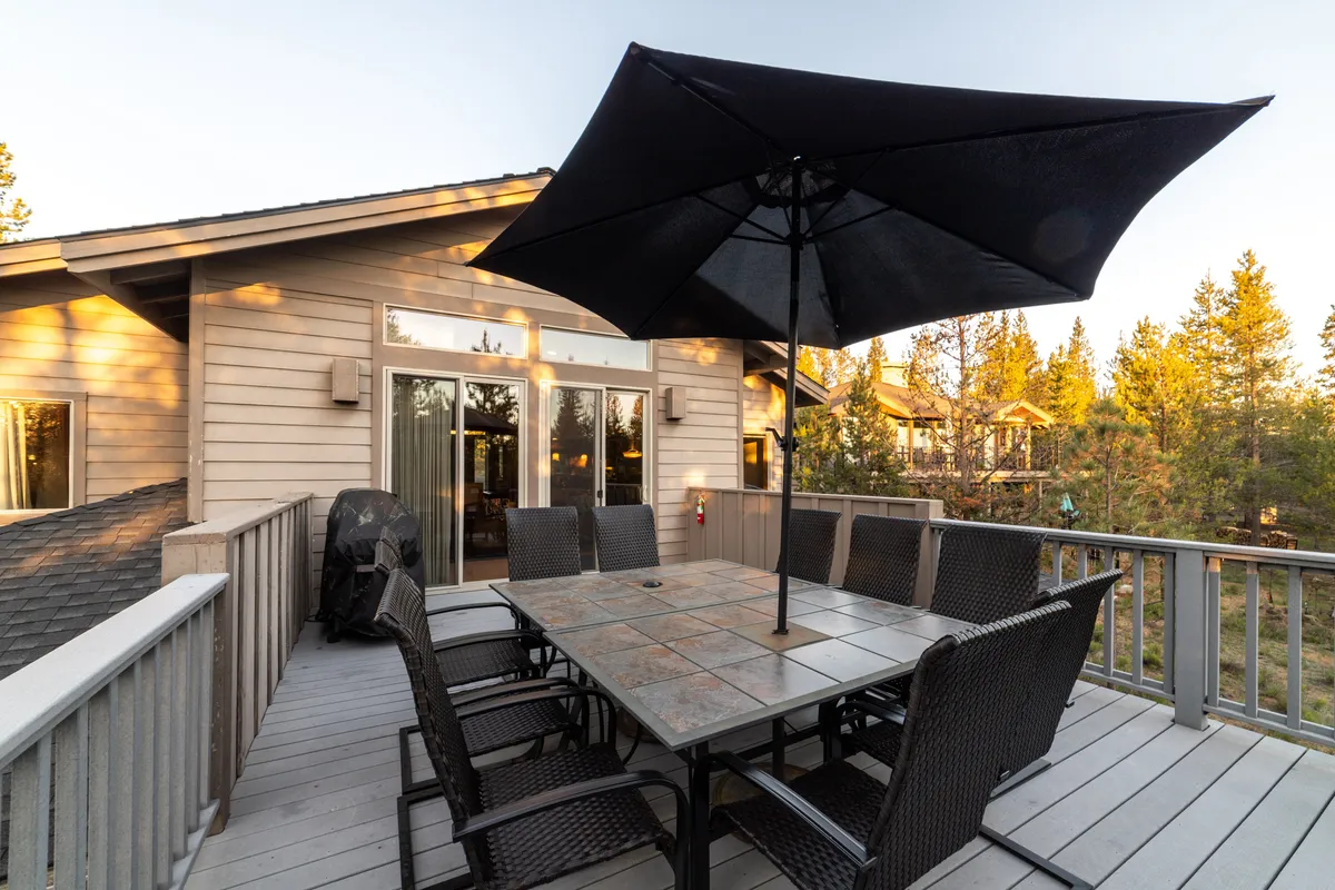 A large outdoor dining table with a black umbrella sits atop a wooden deck, framed by trees and natural surroundings. The deck hints at outdoor relaxation and social gatherings, with space for guests to unwind after a day of recreation.