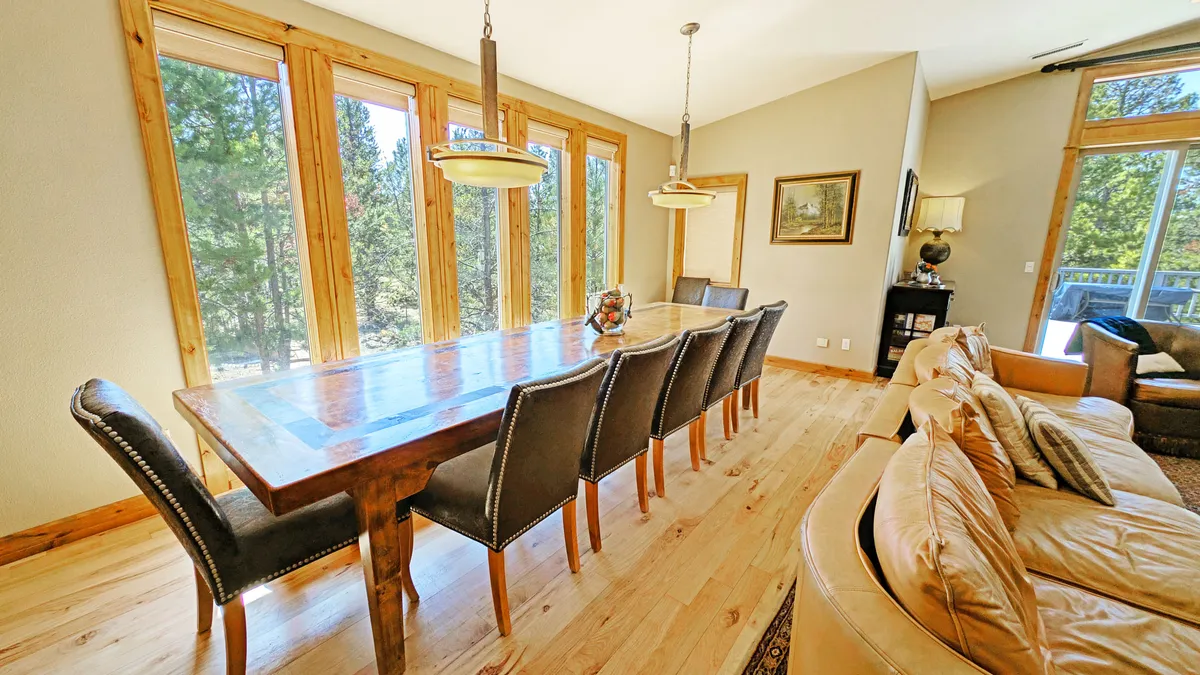 A bright and welcoming dining area with a large wooden table surrounded by elegant chairs, set before expansive windows offering views of the surrounding forest. A comfortable sofa in the background hints at the room’s open layout and connection to other living spaces.