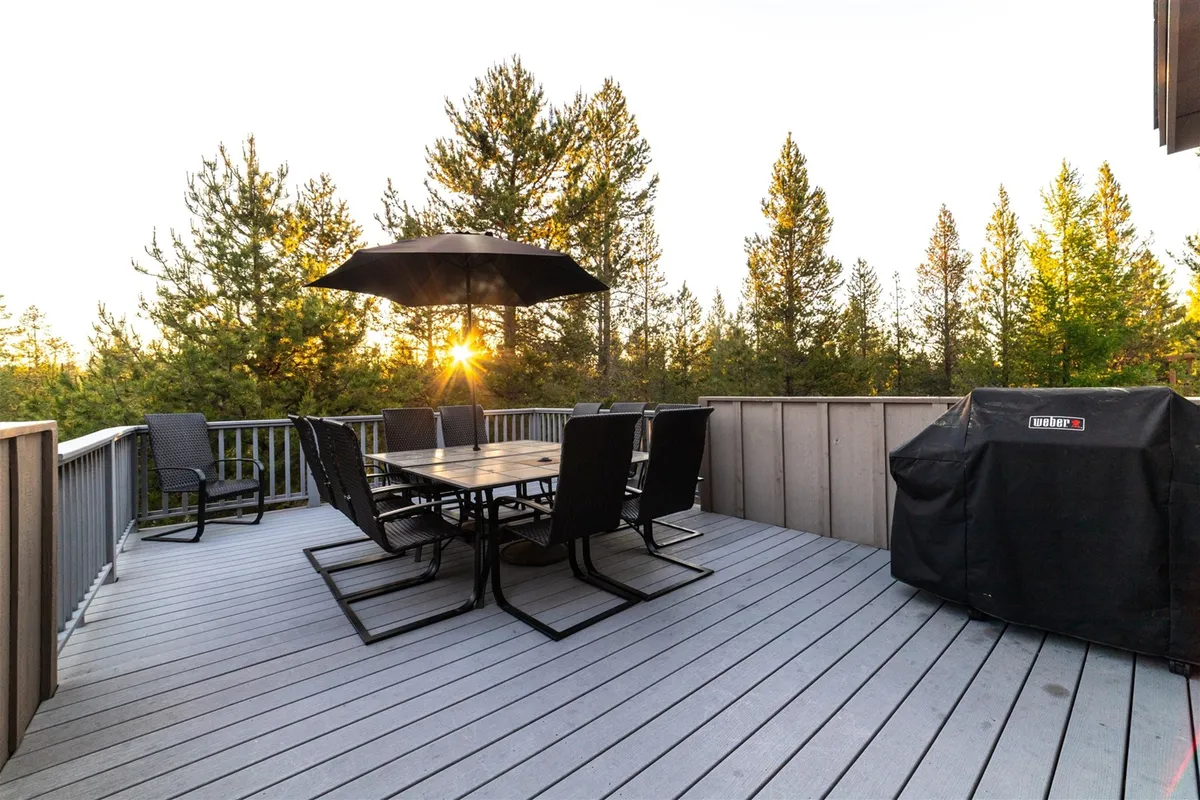 An upper deck scene featuring a large dining table set under an umbrella, a gleaming stainless Weber grill, and a backdrop of lush pine trees. The setting is perfect for outdoor dining, barbecuing, and enjoying nature.
