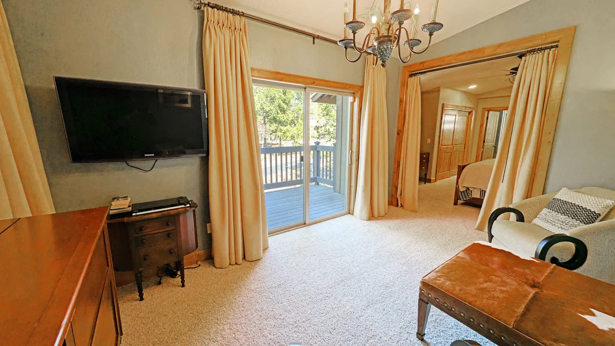 A warm and welcoming living space with a sliding glass door that opens directly to the deck, allowing for easy indoor-outdoor flow. The interior includes a stylish armchair, a coffee table with decorative accents, a wall-mounted TV, and a chandelier, with natural light streaming in from the deck entrance.