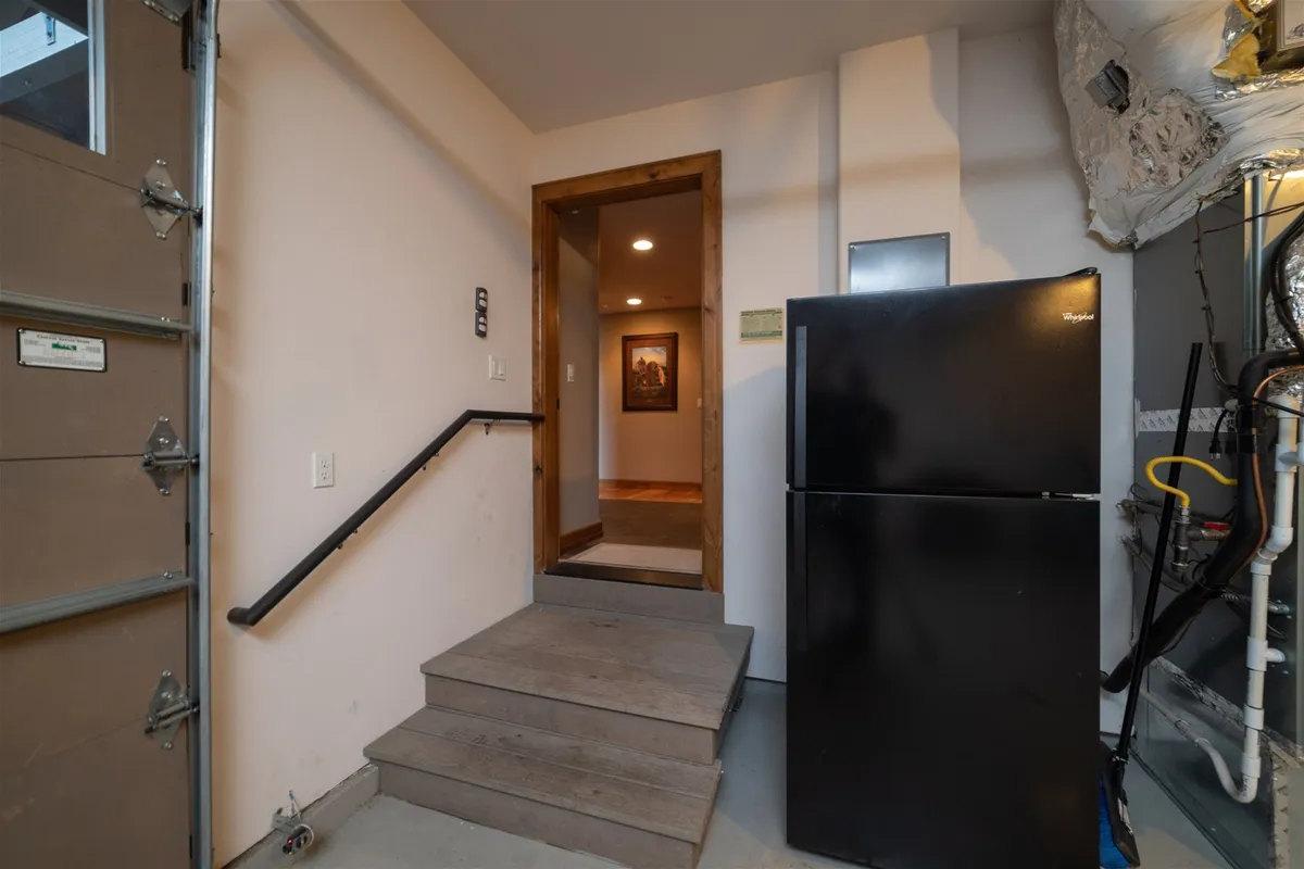 This image features a black refrigerator set against a light-walled space, with a staircase and a doorway leading to another room. The view suggests a transition area, possibly a mini-kitchen or auxiliary space, signifying dual-level access and secondary kitchen amenities.