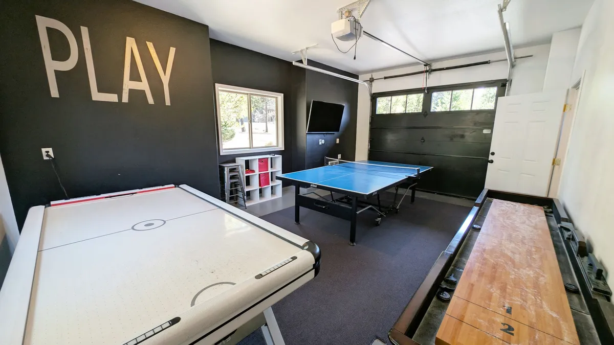 The photo features another angle of the same game room, with the air hockey table and ping pong table front and center, as well as the sliding garage door and a large window bringing in natural light. The layout encourages group activities and socializing.